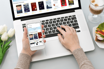 Person's hand holding a phone with Amazon Prime Reading on the screen about a laptop