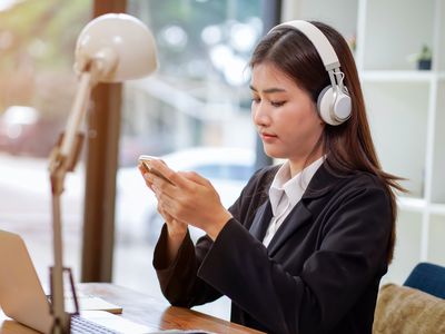 Person wearing headphones looking at smartphone in front of laptop