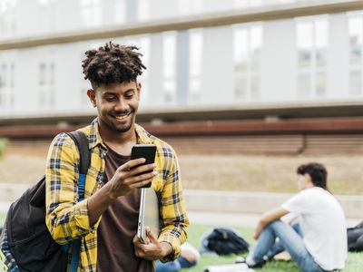 A college student on college campus walking and looking at a smart phone and holding a laptop