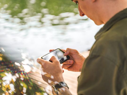 A man in a green shirt playing Pokemon Unite on his iPhone outside near a lake.