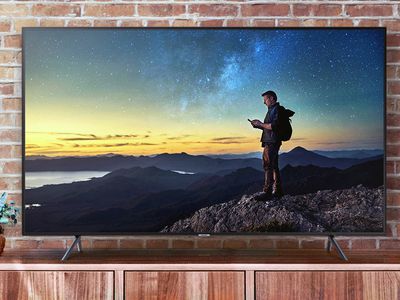 A Samsung NU7100 Series UHD TV in front of a brick wall. The TV is on a media cabinet and is displaying a picture of a man who is backpacking and has reached the top of the mountain's summit. There is a beautiful view but he is looking at his smartphone.