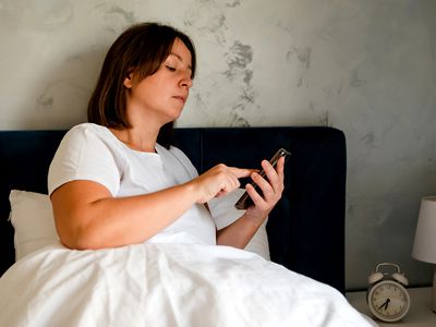 A woman wearing a white T-shirt sitting in bed holding an iPhone 14 smartphone. 