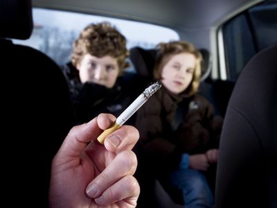 Hand holding a burning cigarette in foreground of car interior with two small children in the backseat