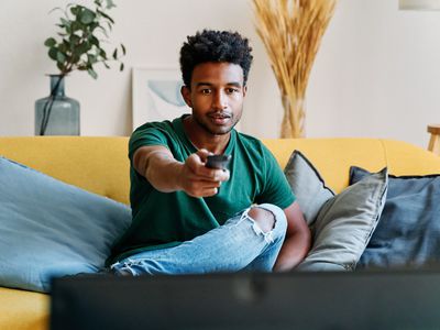 A man in a green shirt and blue jeans sitting on a yellow couch. There are three blue throw pillows on the couch. He is using a typical TV remote.