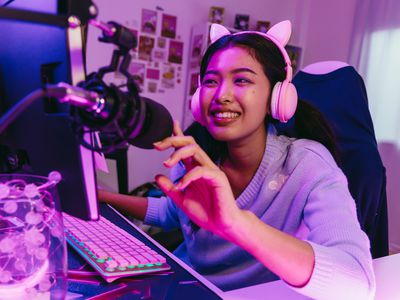 A woman sitting at her desk smiling broadly at a PC monitor while wearing headphones with ears.