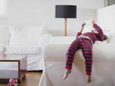 Child in pajamas lying on couch armrest with mobile device in two hands.