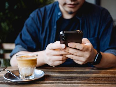 A man sitting at a table in a cafe with a latte on it while checking Snapchat on his black iPhone smartphone.