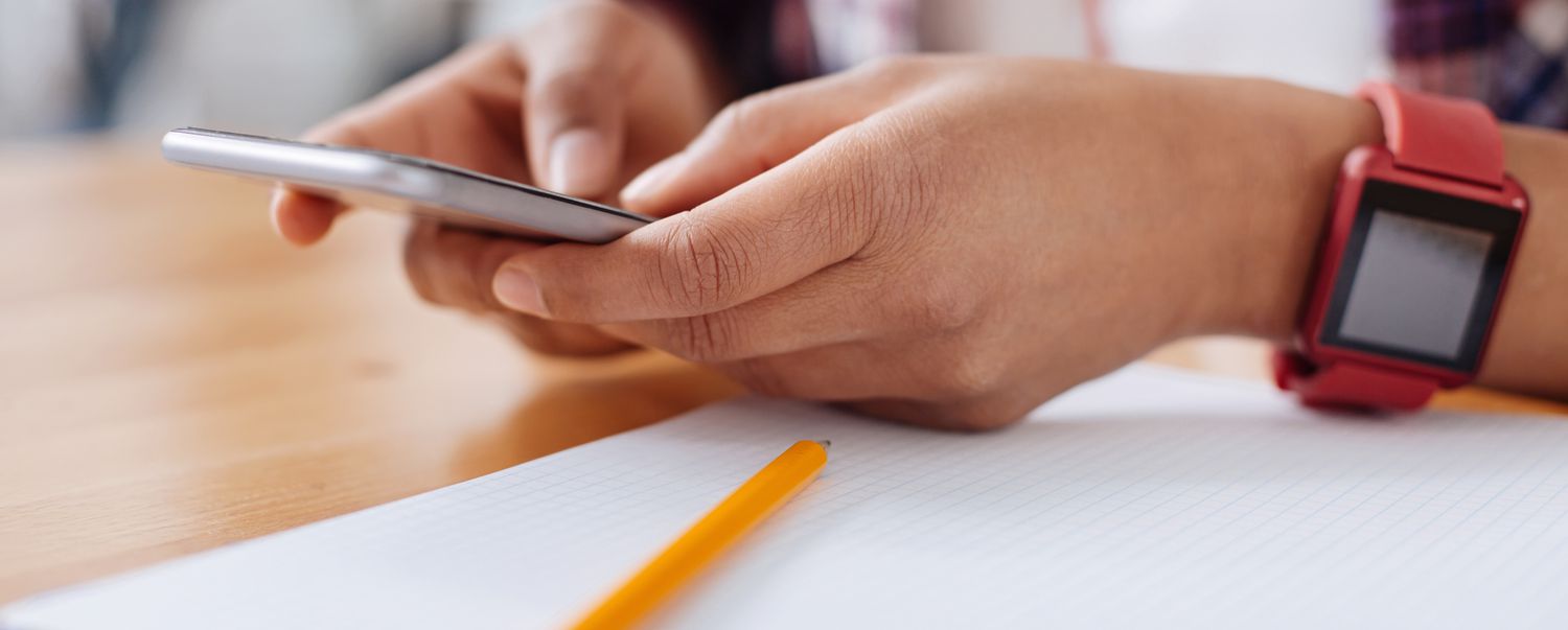 Hands holding a smartphone with a red smartwatch on wrist over a pencil and paper.