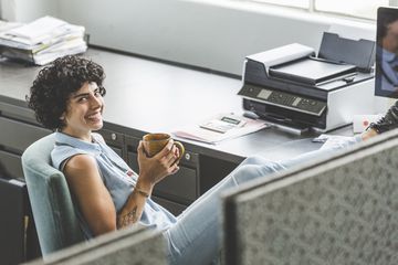 Someone sitting in front of a personal printer with their feet on the desk and cup of coffee in their hands.