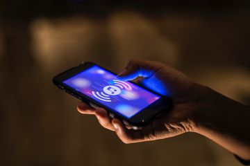 A stock, close-up photo of a person's hand holding a smartphone, the screen of which has a music note symbol in the middle with soundwave marks surrounding it, on a purple and pink background.