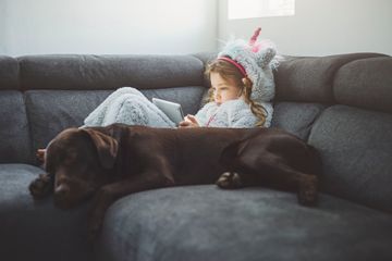 Young girl in a unicorn onesie using tablet, dog by her side