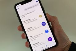 Photo of hand holding iPhone XS Max with screenshot of Google Assistant actions listed (e.g., Get good news, Topical news, Help, etc.)