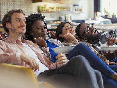 Four people sitting on a couch eating popcorn and watching tv.