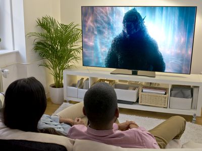 A man and woman sitting on a couch in a brightly lit lounge room watching Godzilla in Monarch Legacy of Monsters on their TV.