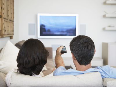 Two people sitting on a couch in front of a televisions. One holds a remote pointed at the television.