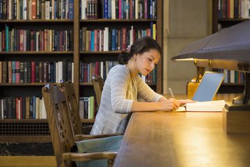 Woman using a Laptop in a Library working on a Research Paper on long wooden table