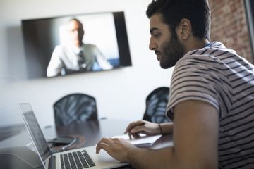 Man sat looking at a laptop while a wall-mounted TV plays in the background.