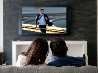 A man and a woman sitting on a couch watching Mission: Impossible Dead Reckoning on a TV hung on a wall.