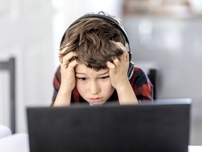 A boy wearing headphones sitting at a black laptop computer looking sad and stressed.