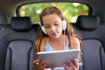 Shot of a young girl sitting in a car wearing headphones and using a digital tablet.