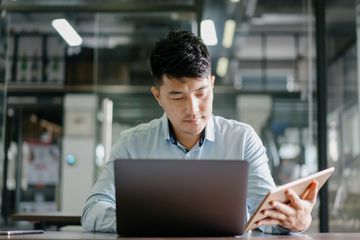 A man sitting at a desk looking at a laptop and tablet at the same time