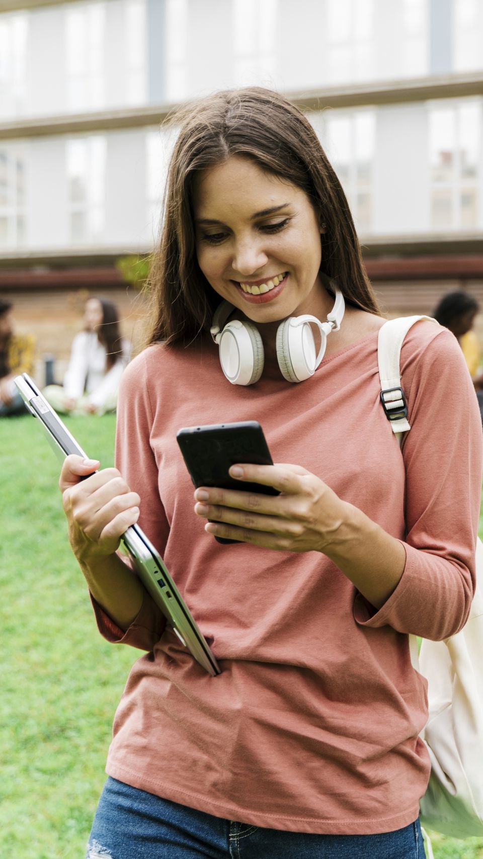 Female student walking with a tablet, smartphone, and headphones.