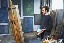 Male artist holding paint brush looking at canvas on easel in studio