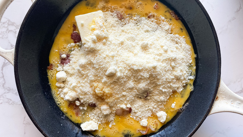 Eggs, cooked bacon, butter, and grated parmesan cheese in a skillet.