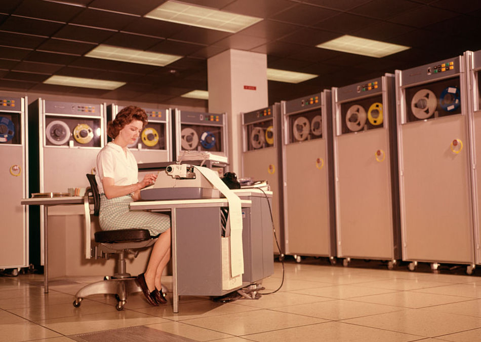 A woman enters data into a 1960s B-5000 computer surrounded by reels of magnetic tape units.