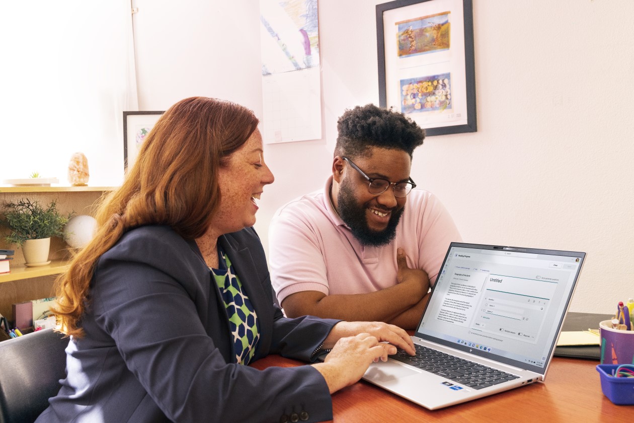 Decorative. Two educators sitting at a table in a classroom and working together on a laptop.