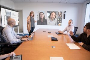 Two woman and three men (one in a wheelchair) are hosting a coordinated meeting in a large conference room and are joined by several virtual attendees on a projection screen in Front of Room view. The in-person team is interacting with remote attendees through chat and live reactions while following the woman at the Surface Hub. One woman is on an iPad logged into the Teams meeting, two men joining the meeting from an iPad and Surface.