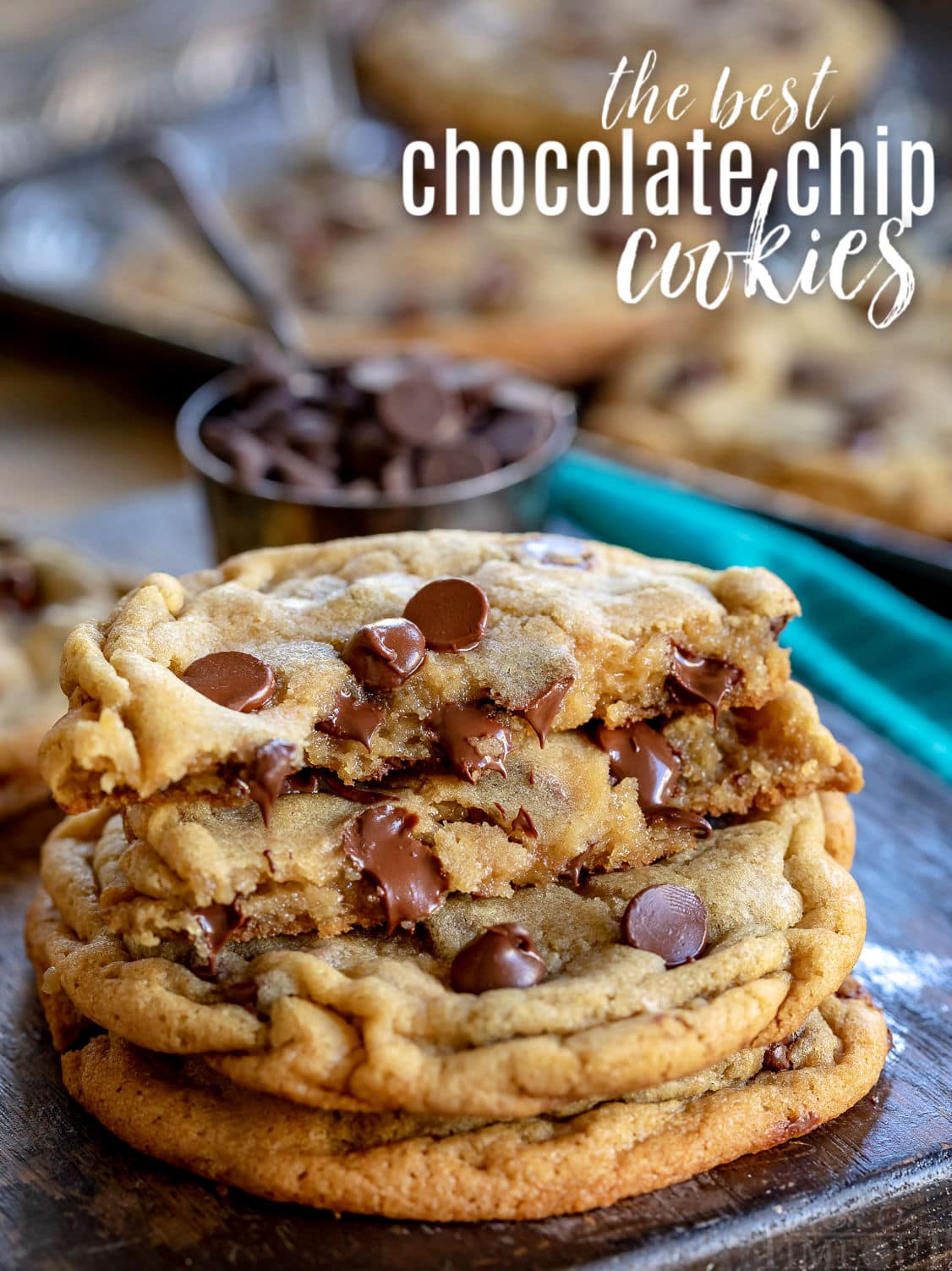 three chocolate chip cookies stacked on dark cutting board with baking sheet in background