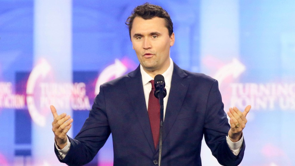 Charlie Kirk, a white man with brown hair, in a suit, gesturing onstage while speaking. The logo "turning point USA is in the background."