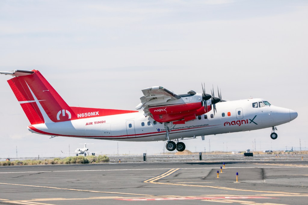 A four-engine turboprop aircraft in a red and white livery takes off from a runway on its way to be modified into a hybrid electric aircraft. Other airplanes can be seen in the distance.