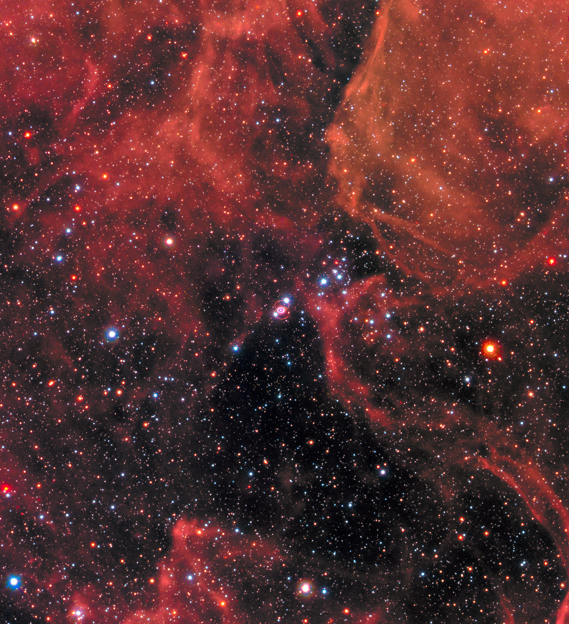 Clouds of gas and dust in shades of red cover the top of the image and a little bit of the bottom. Right in the middle is Supernova 1987a, the remnant of an exploded star. The site of the supernova is surrounded by an illuminated pinkish ring. Two faint outer rings are also visible. Stars and galaxies shine throughout, giving off red and blue light.