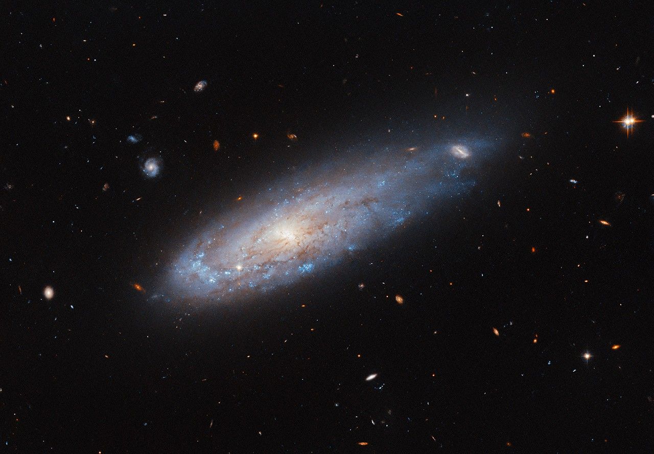 A spiral galaxy with a disk that glows visibly from the center. It has faint dust threaded through it. A spiral arm curves around the left edge of the disk and is noticeably denser with bright blue spots that hold hot and new stars. On the opposite side, the disk stretches out into a short tail where it covers a distant background galaxy. Other distant galaxies and some nearby stars are visible are also visible in this image.