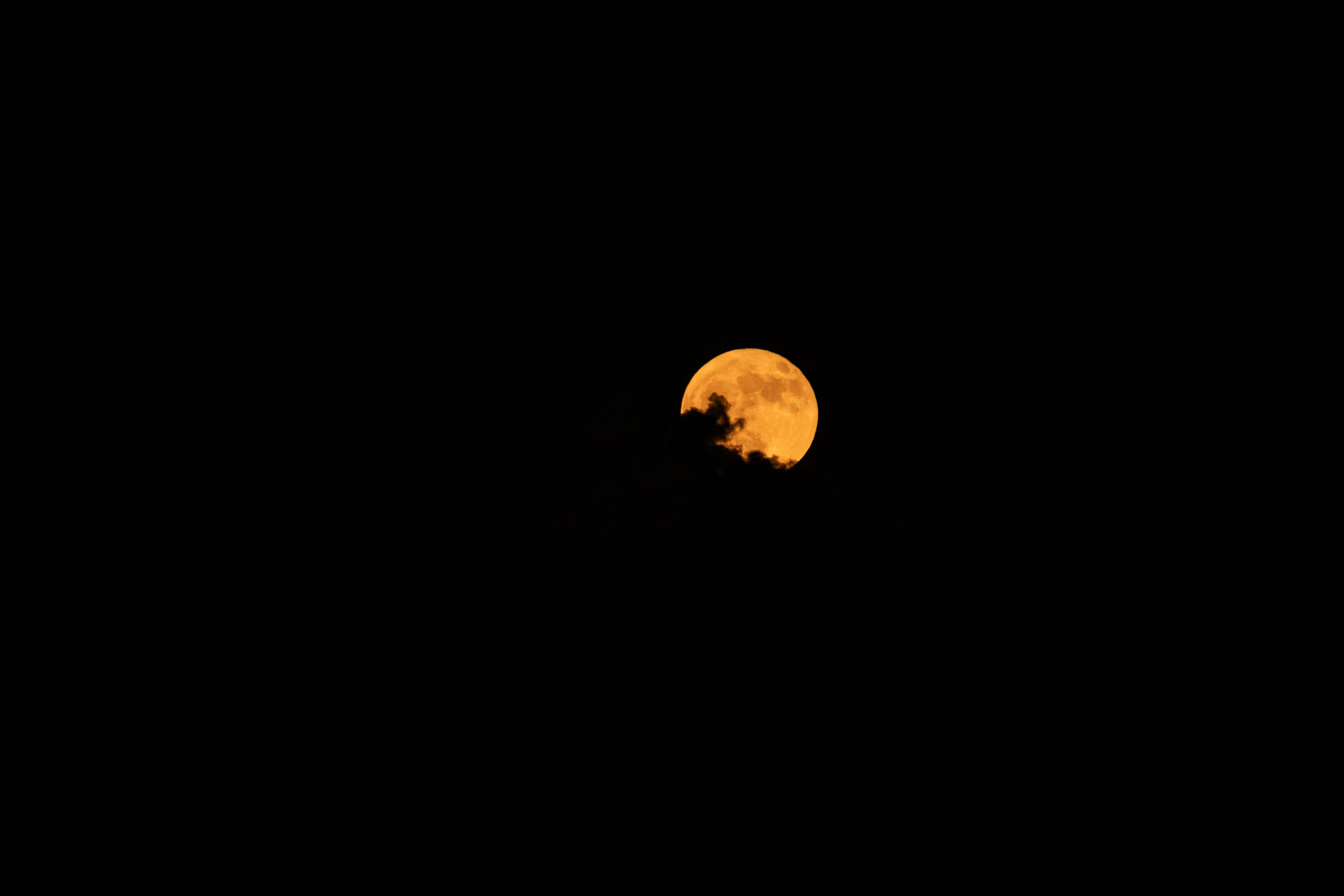 An orange full moon is the only thing you can see in this image. The sky is black, and the outline of some clouds are visible against the Moon.