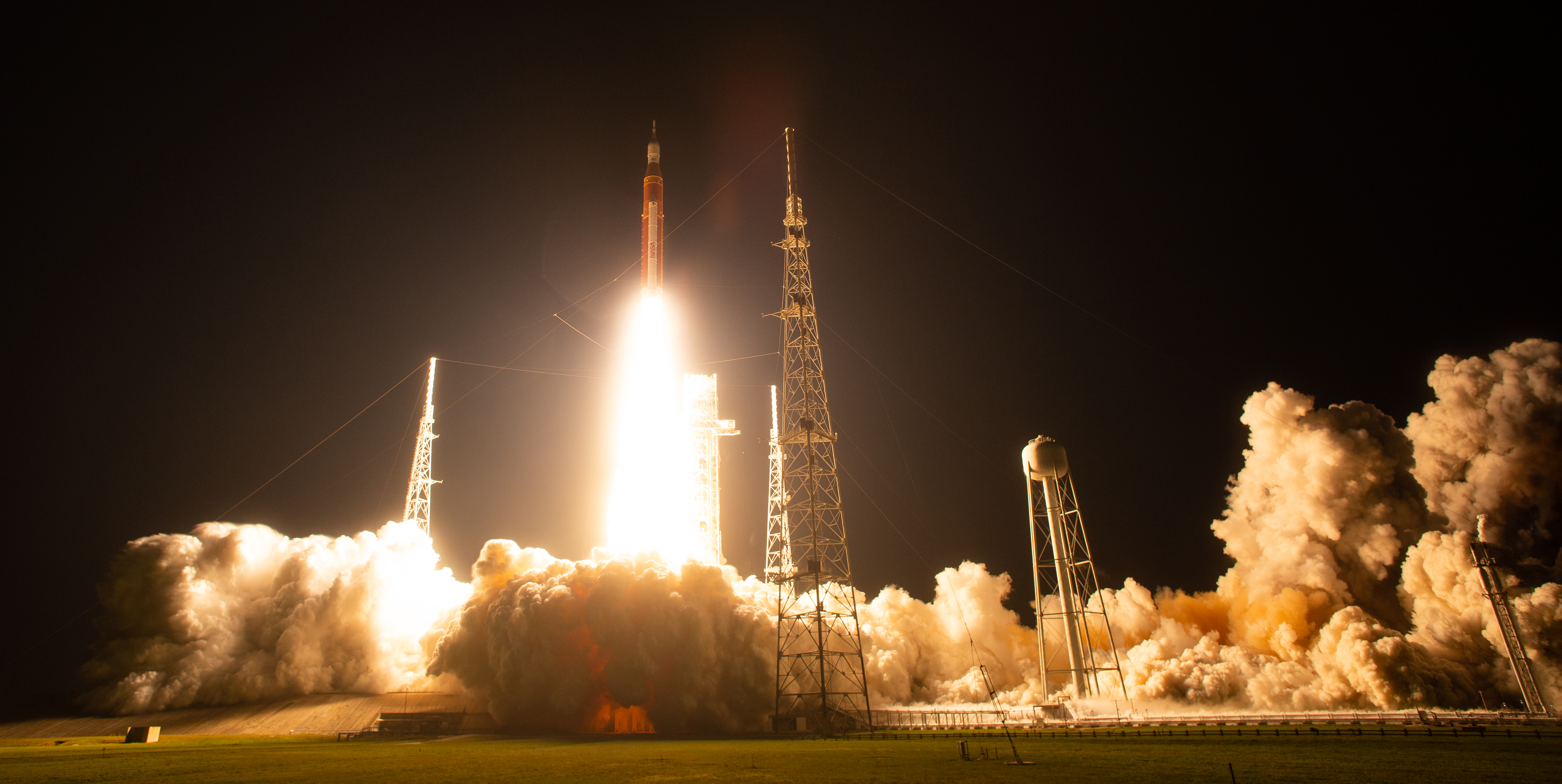 An orange rocket lifts off from a launchpad. The sky is black, but the fire coming from the engines lights up the surrounding area, highlighting clouds of white vapor spreading across the ground.