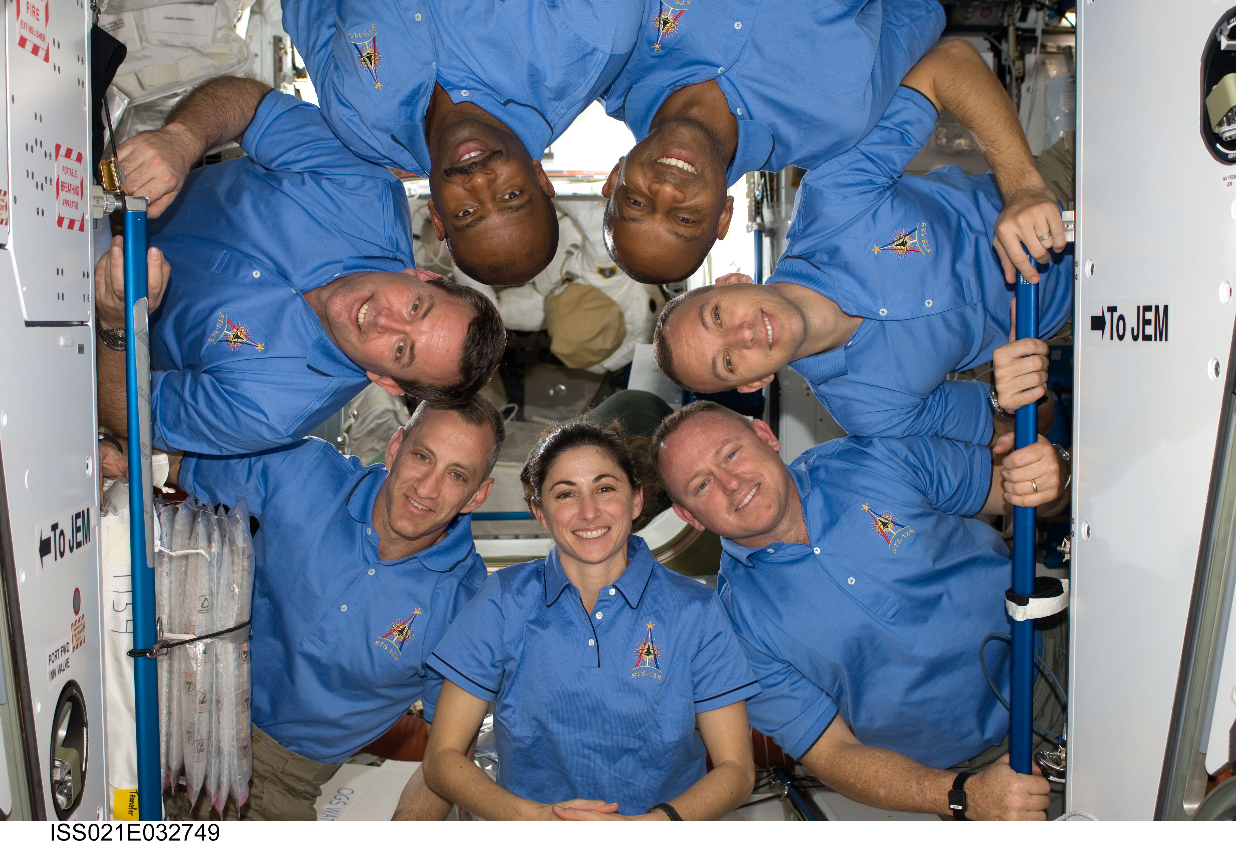 Seven astronauts pose for a photo on the International Space Station. They are arranged with their heads in a circle, with a few people upside down. They're all wearing blue polo shirts with an STS-129 logo on them.