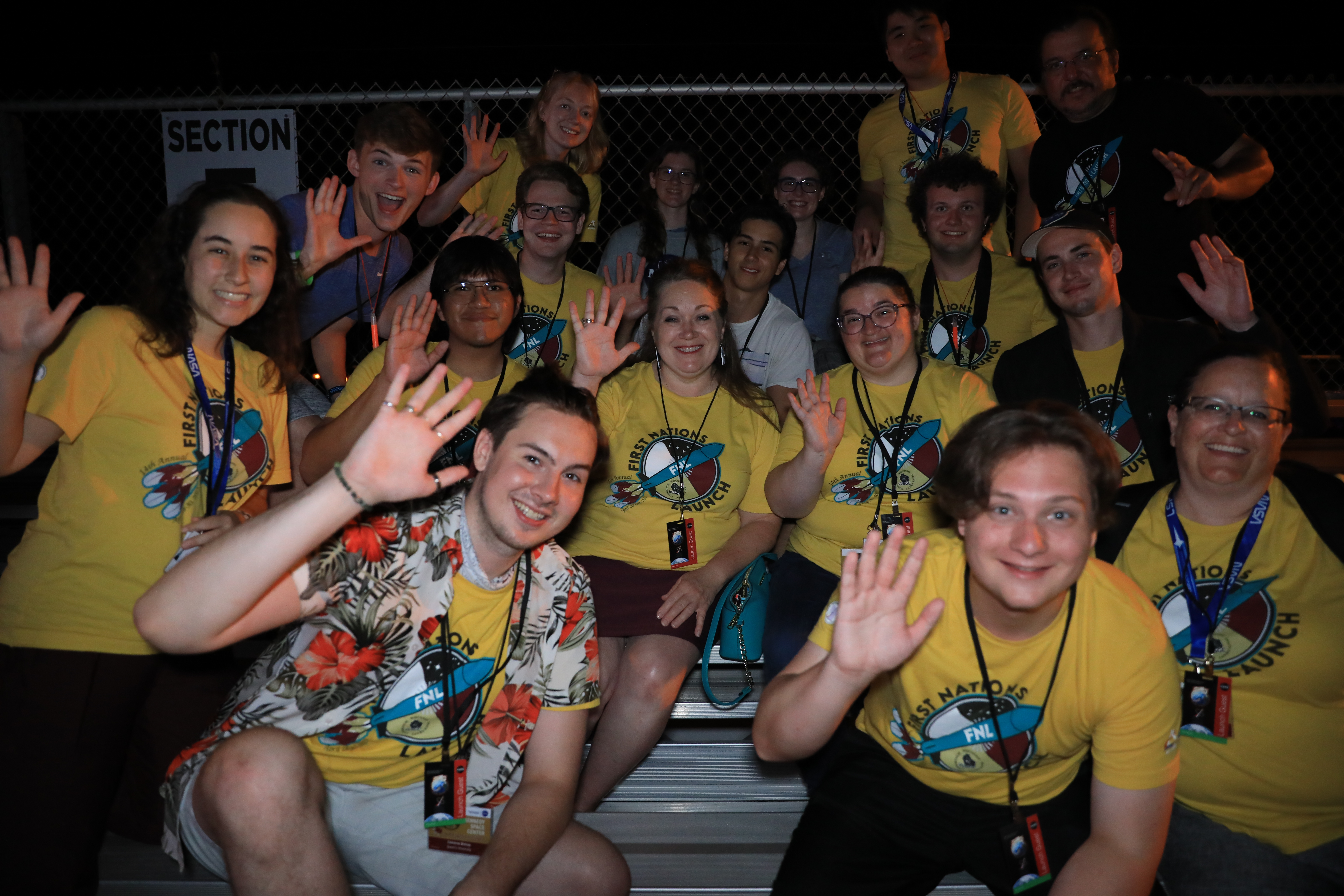 A group of people wearing yellow t-shirts wave at the camera as they sit outside at night. They are members or advisors of winning teams from the 14th First Nations Launch High-Power Rocket Competition. The competition comprises students from tribal colleges and universities, Native American-Serving Nontribal Institutions, and collegiate chapters of the American Indian Science and Engineering Society.