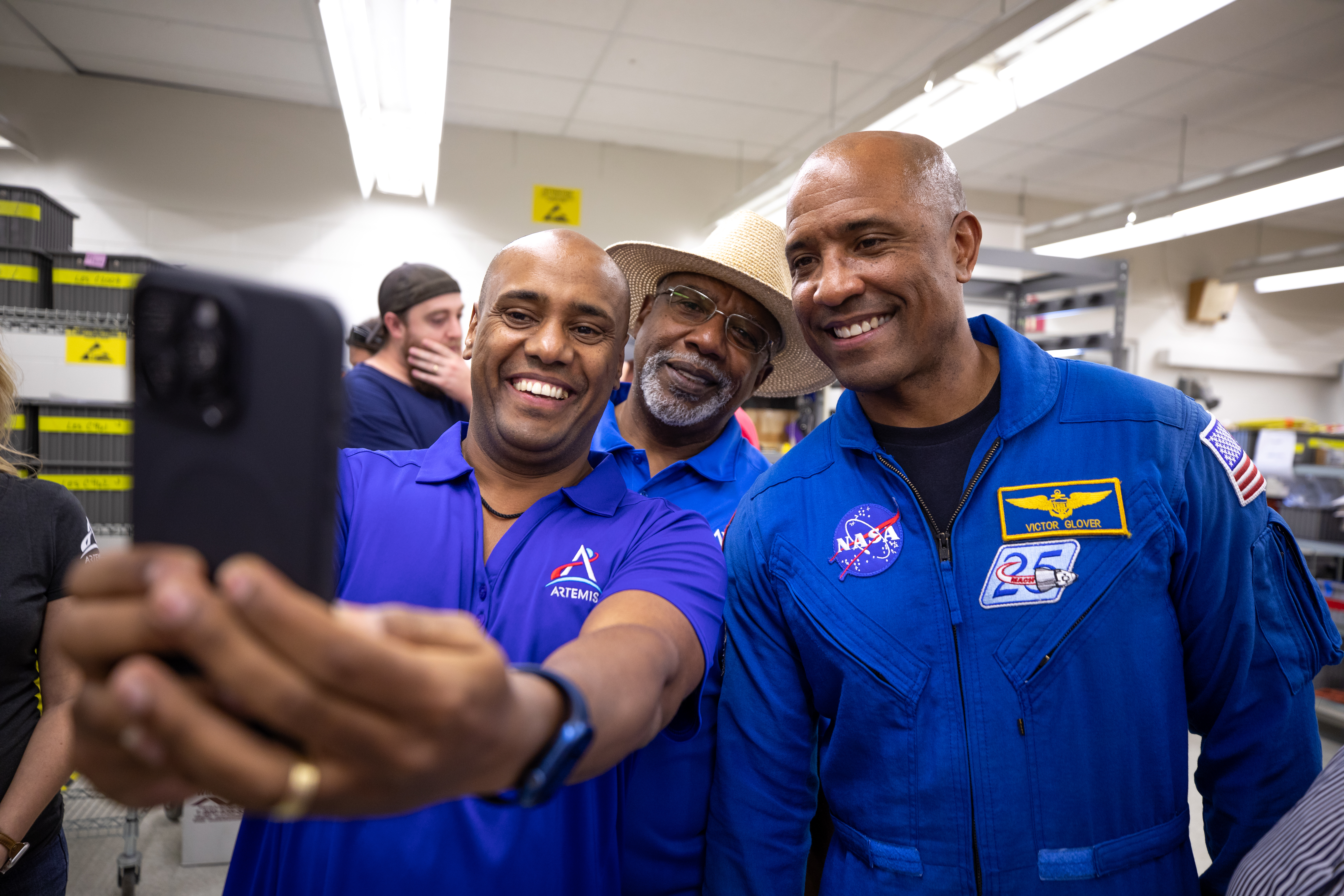 Three African American men - two Kennedy Space Center employees wearing blue polo shirts with an Artemis logo (left, middle back) and NASA astronaut Victor Glover (right) - pose for a photo taken with a cell phone.