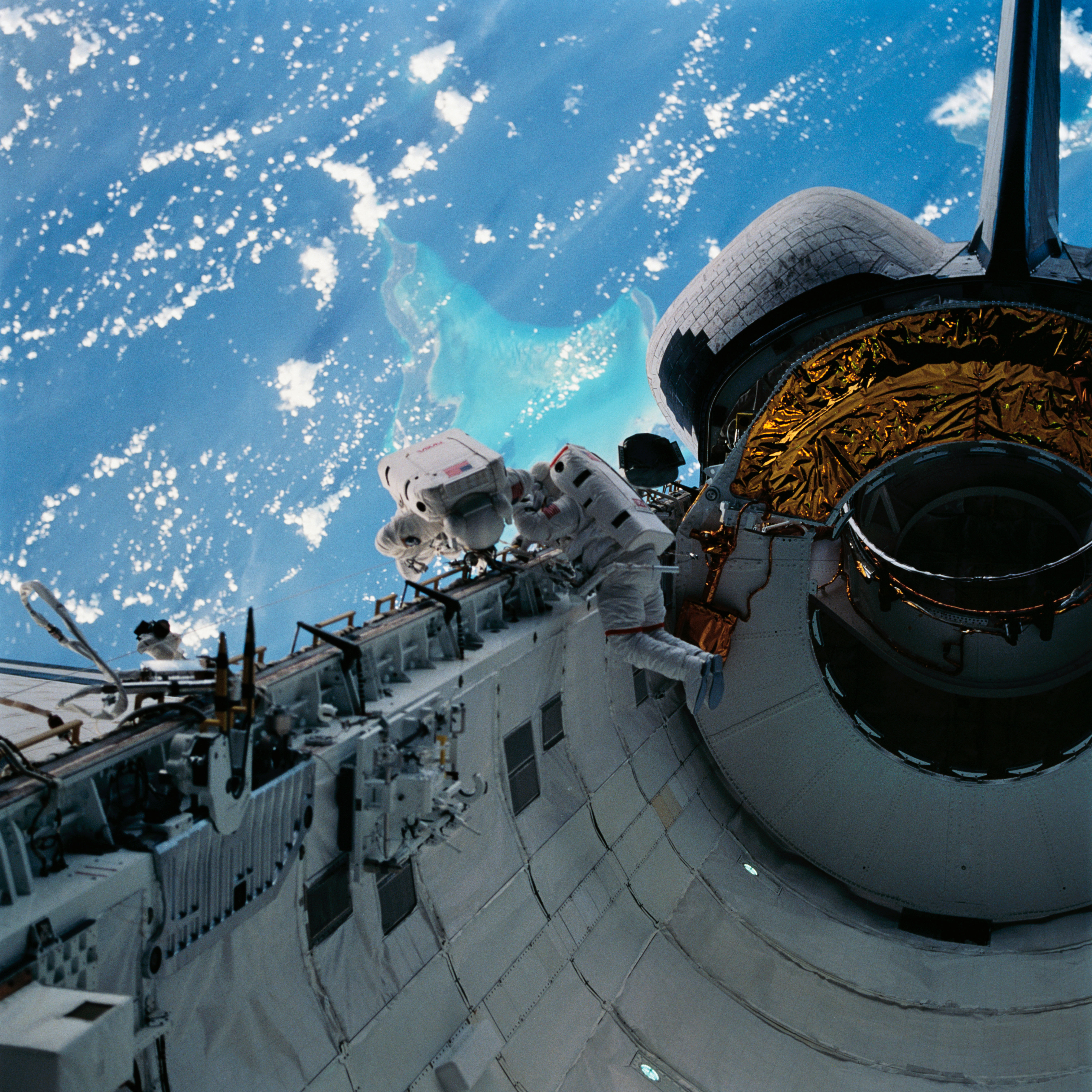 Two astronauts in white spacesuits inspect equipment outside of space shuttle Discovery during a spacewalk. The Caribbean Sea and part of the Bahama Islands chain is visible behind them.