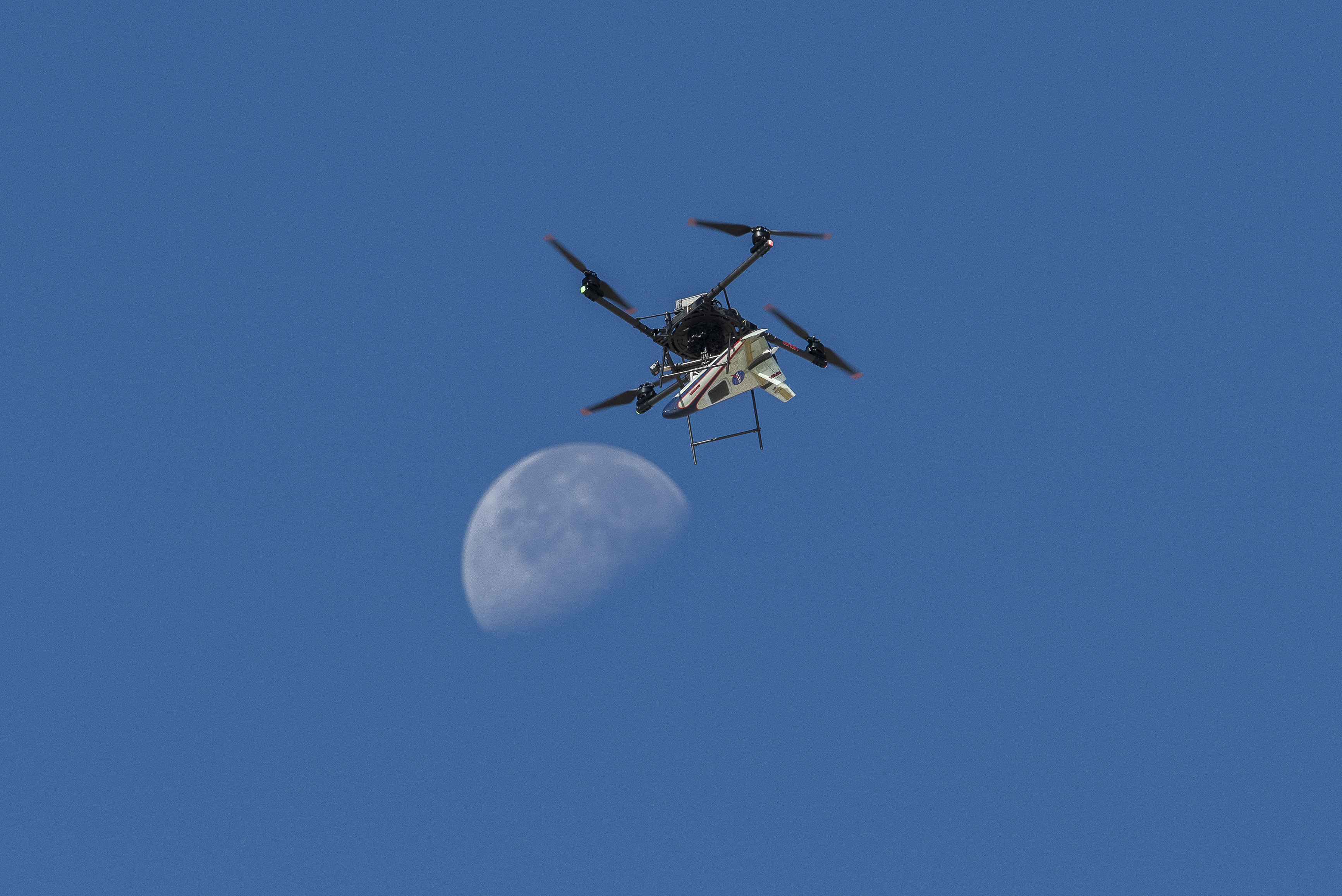 A white, blue, and red probe attached to a rotor with four blades flies in the blue sky, just above the Moon.