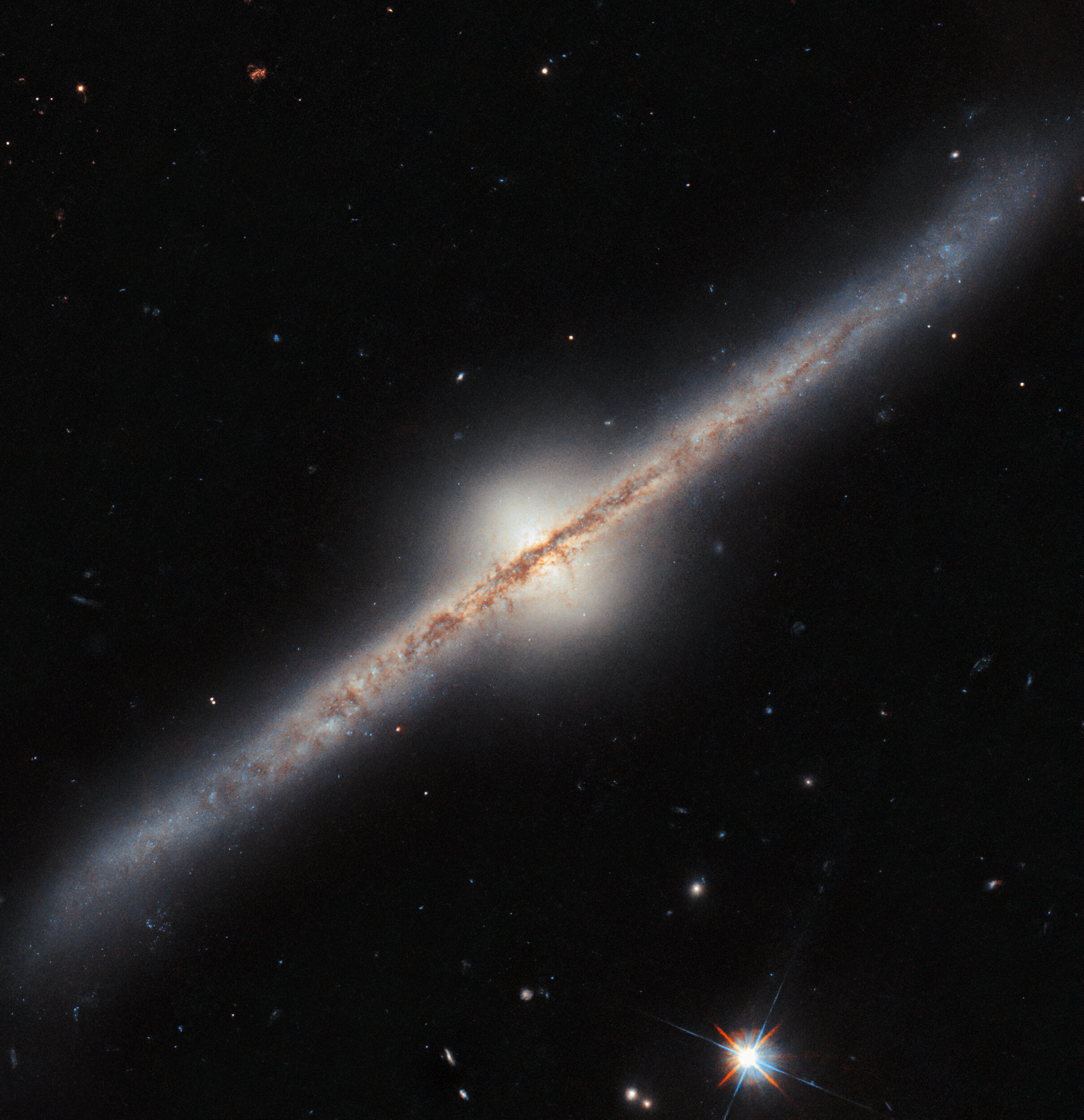 A spiral galaxy seen directly from the side, such that its disk looks like a narrow diagonal band across the image. A band of dark dust covers the disk in the center most of the way out to the ends, and the disk glows around that. In the center of the galaxy, a whitish circle of light bulges out above and below the disk. Each end of the disk curves slightly. The background is black and mostly empty.