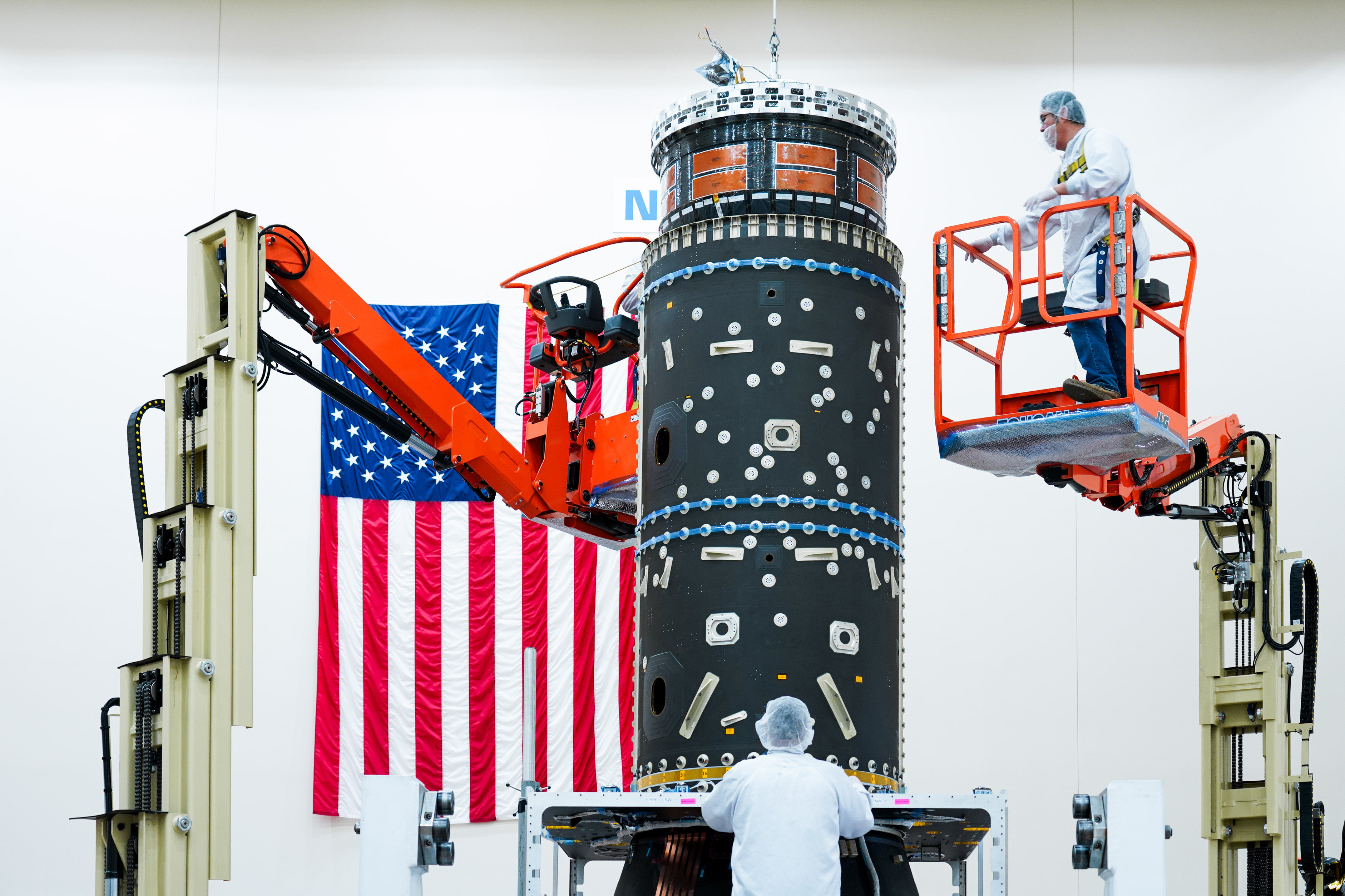 A black piece of equipment with small silver pieces all over it is at the center of this image. This is what houses Gateway's fuel tanks. A person in a blue hairnet, mask, white lab coat, and jeans stands on an orange lift that is several feet in the air. Another person in similar clothing is at the bottom of the piece of equipment at the center of this image. An American flag hangs vertically on the wall.