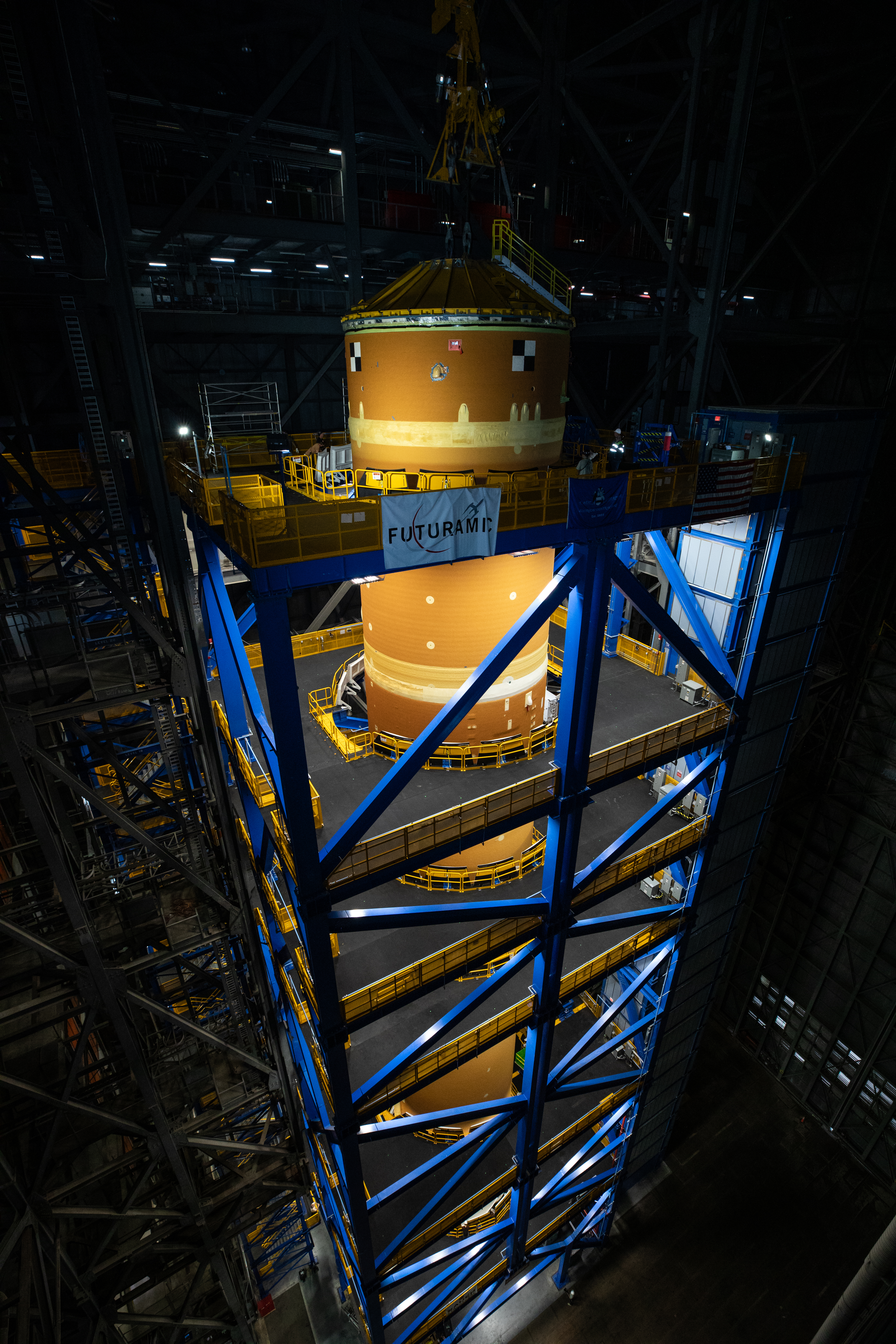 An immense orange cylindrical object - the SLS core stage - is surrounded by a blue metal structure. The space around them is dark.