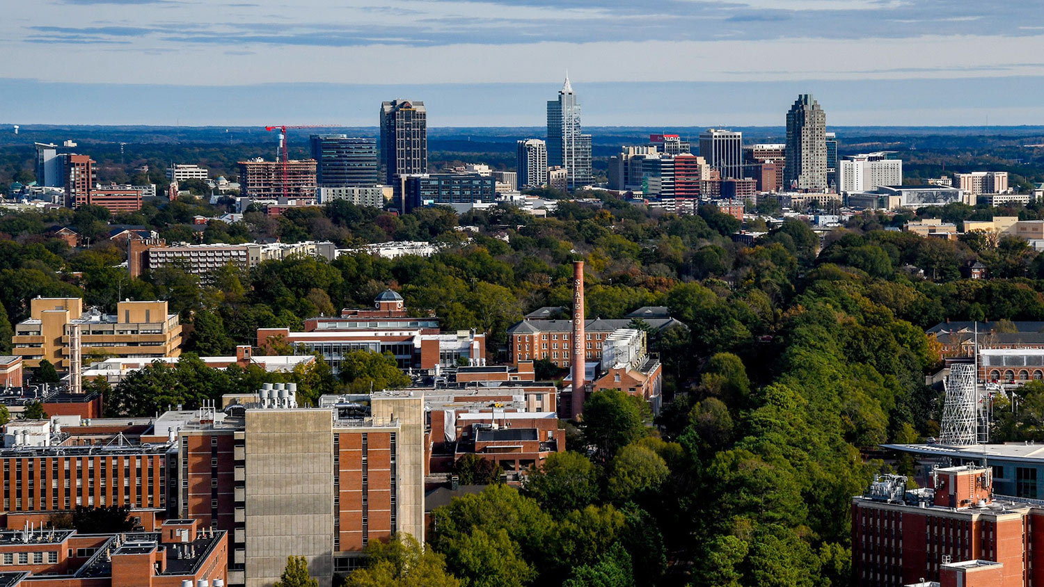 An aerial view of NC State's campus with the Downtown Raleigh skyline behind it.