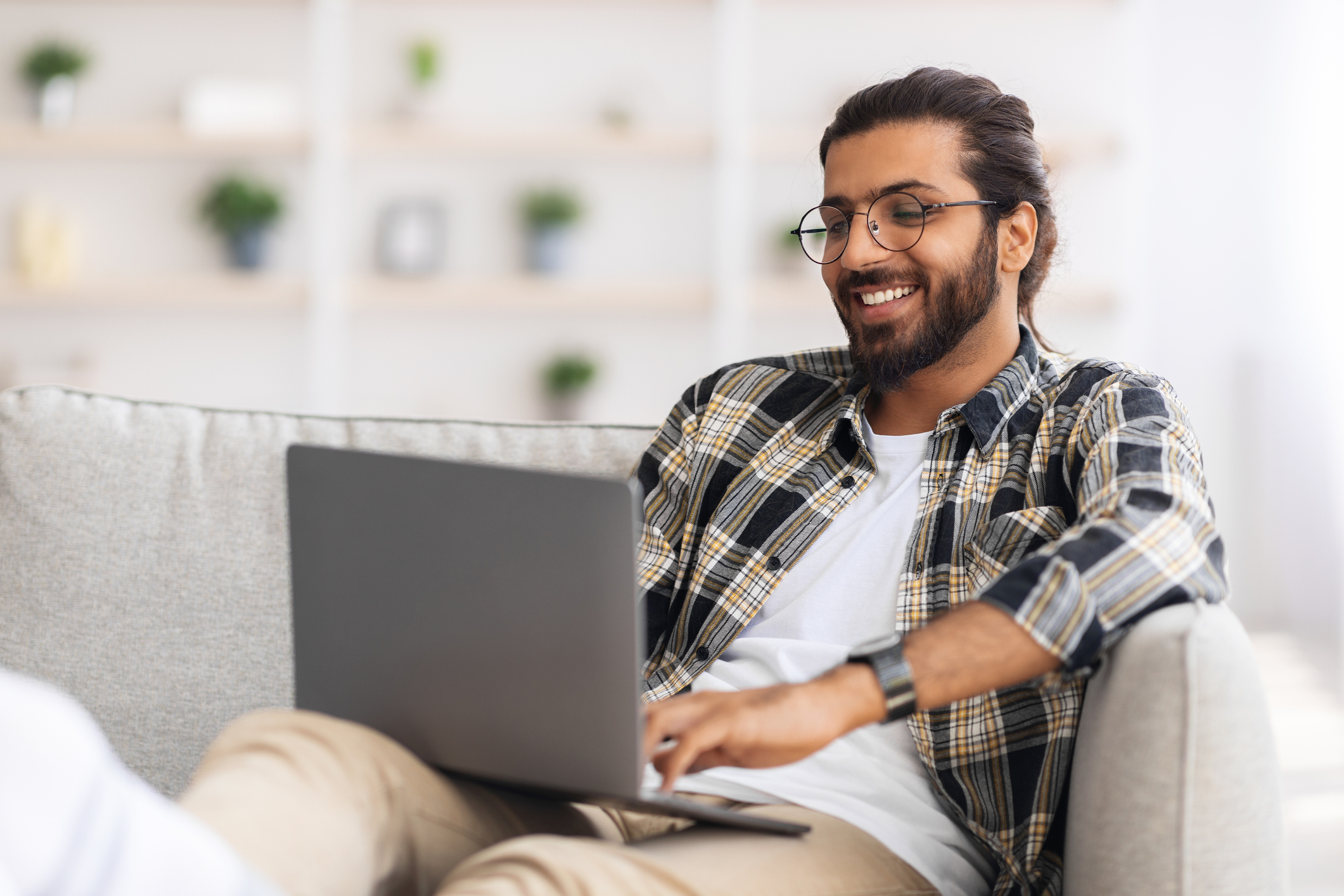 Happy guy with glasses working from home, using modern notebook, sitting on couch in cozy living room