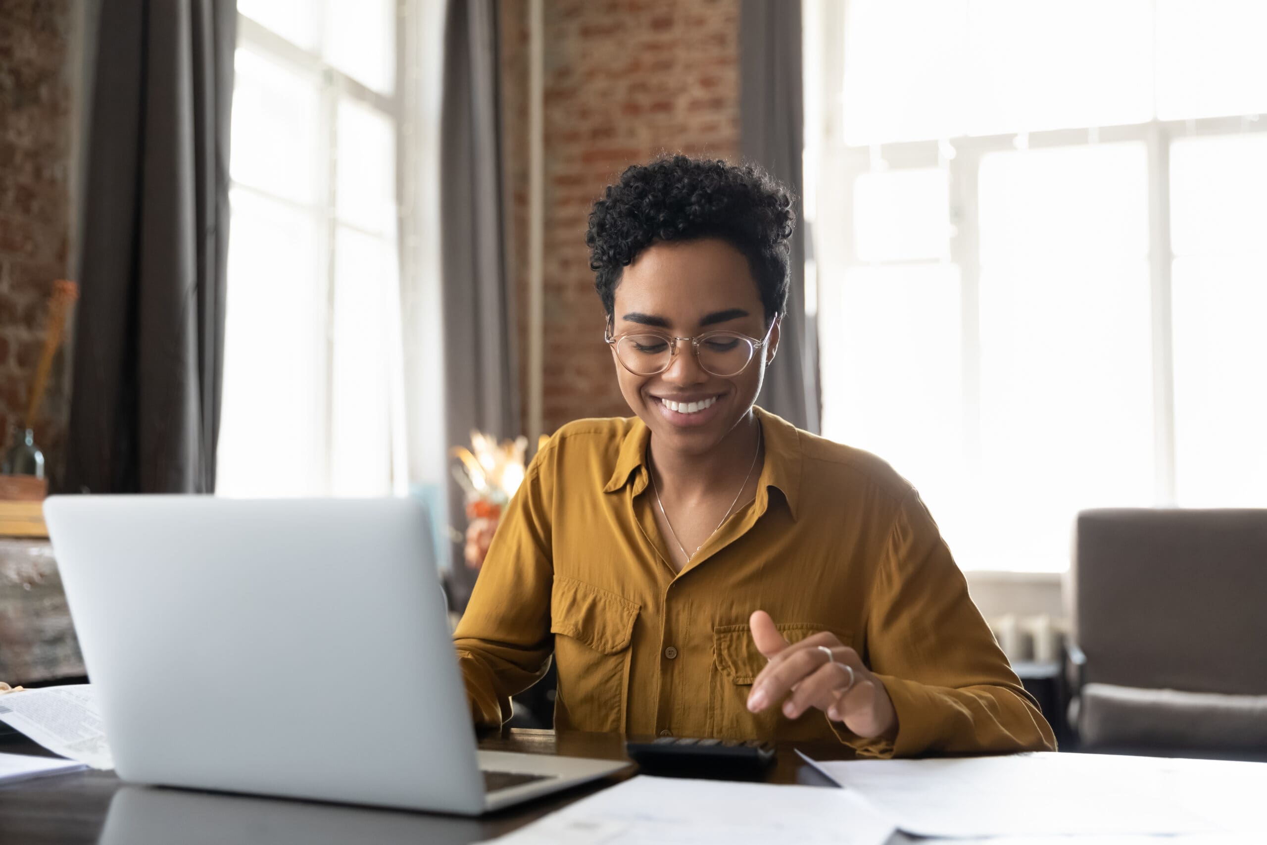 Happy young entrepreneur woman in glasses counting profit, on calculator at laptop computer, analyzing benefits, enjoying financial success, job high result, smiling