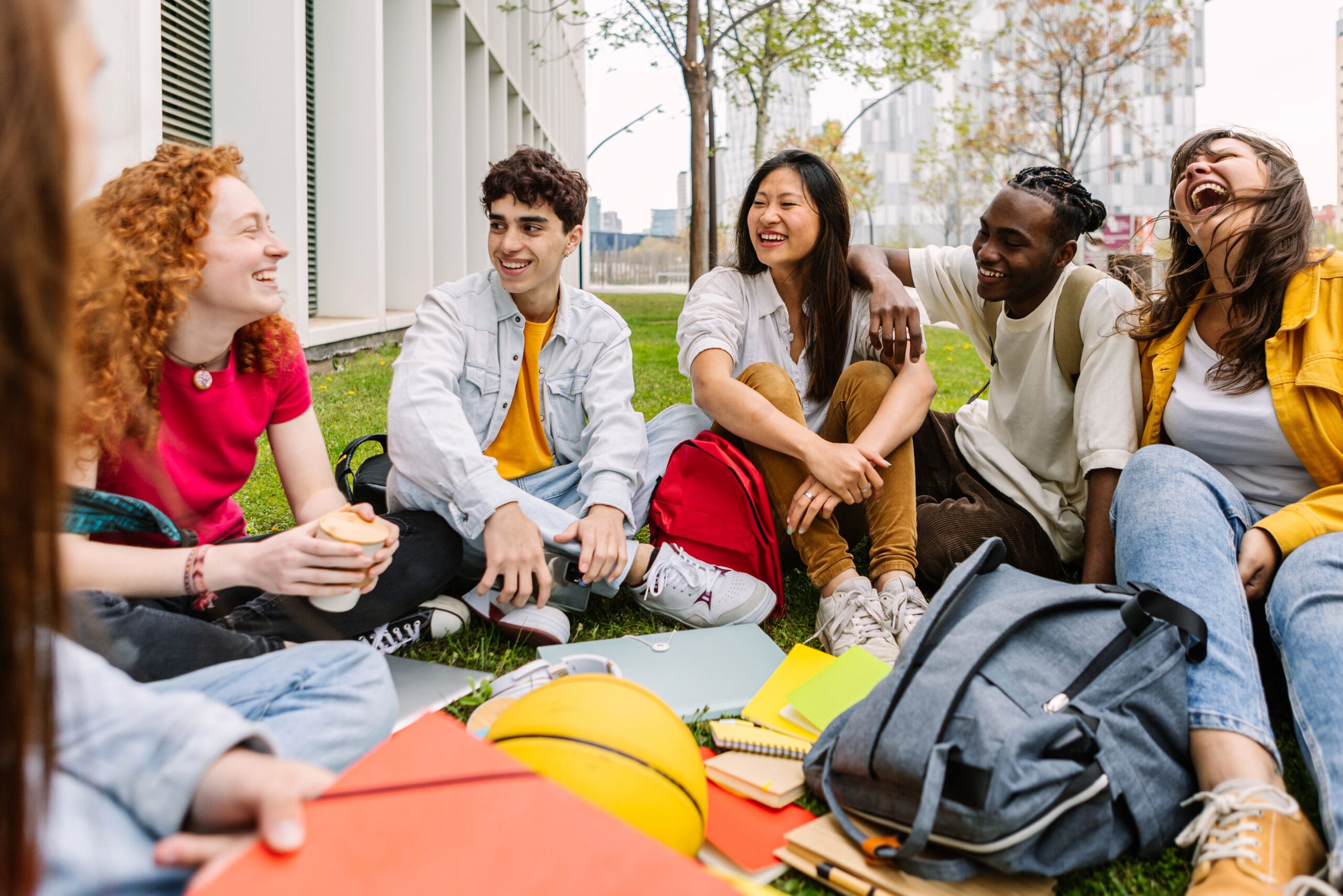 Student friendship concept with multiracial classmate friends sitting together on the grass at campus college park. Millennial people having fun social gathering outside. Youth and education.